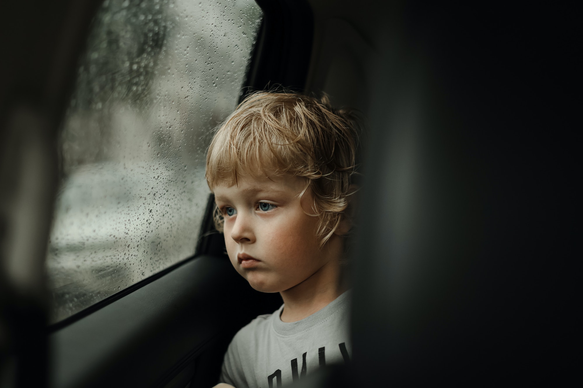 A cute little blond boy looking out of the car window on a rainy day. Bad weather mood, depression N